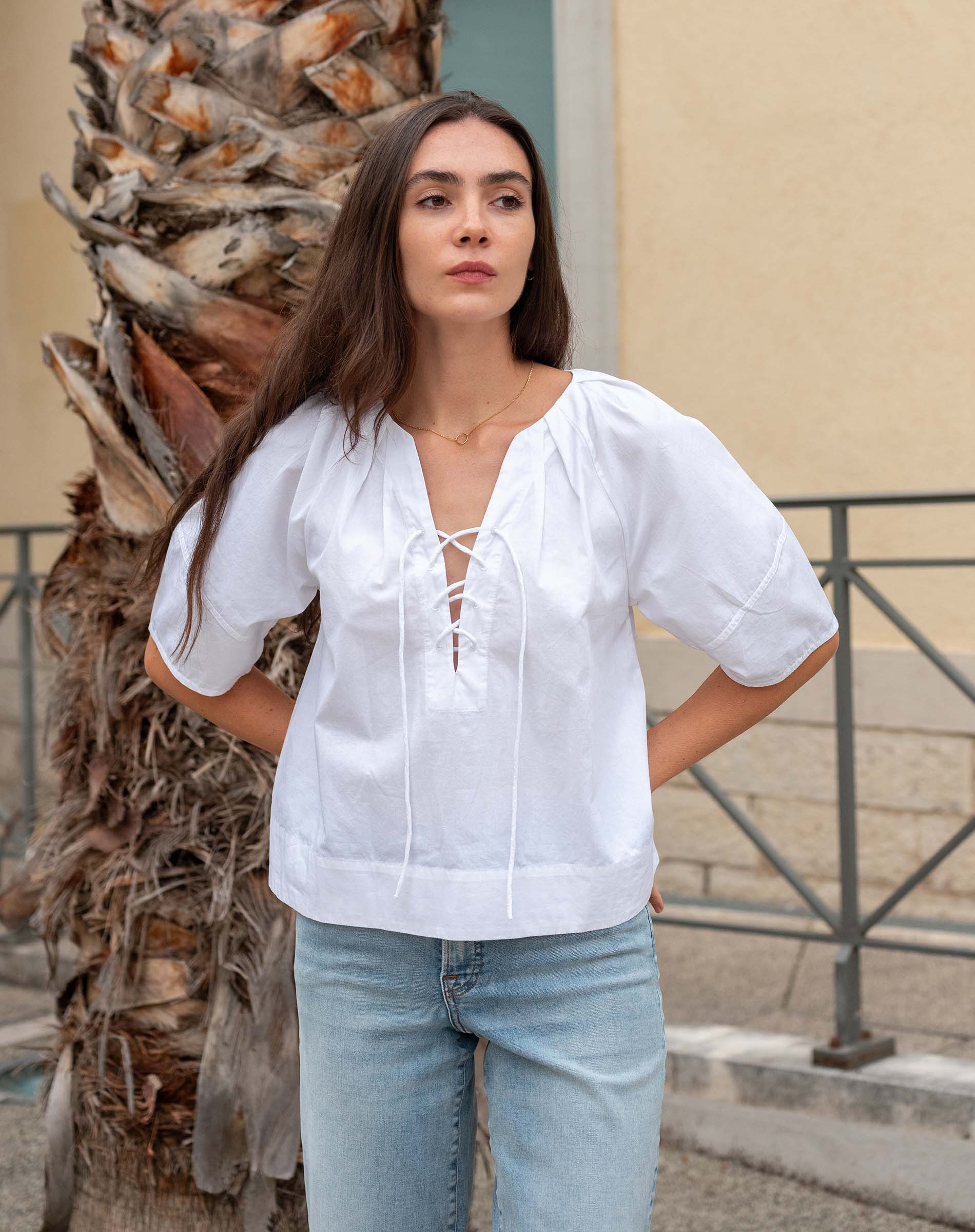 Woman wearing a white blouse and light blue jeans standing next to a palm tree.