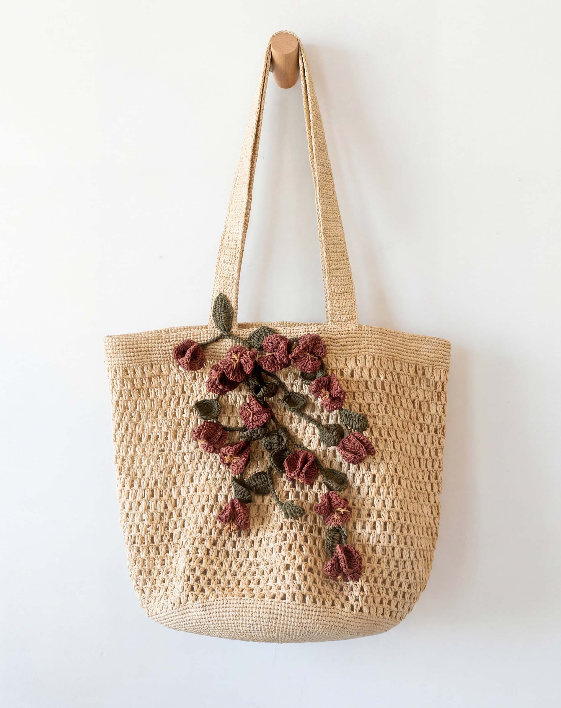 Woven bag with floral embroidery hanging on a wooden hook against a white wall.