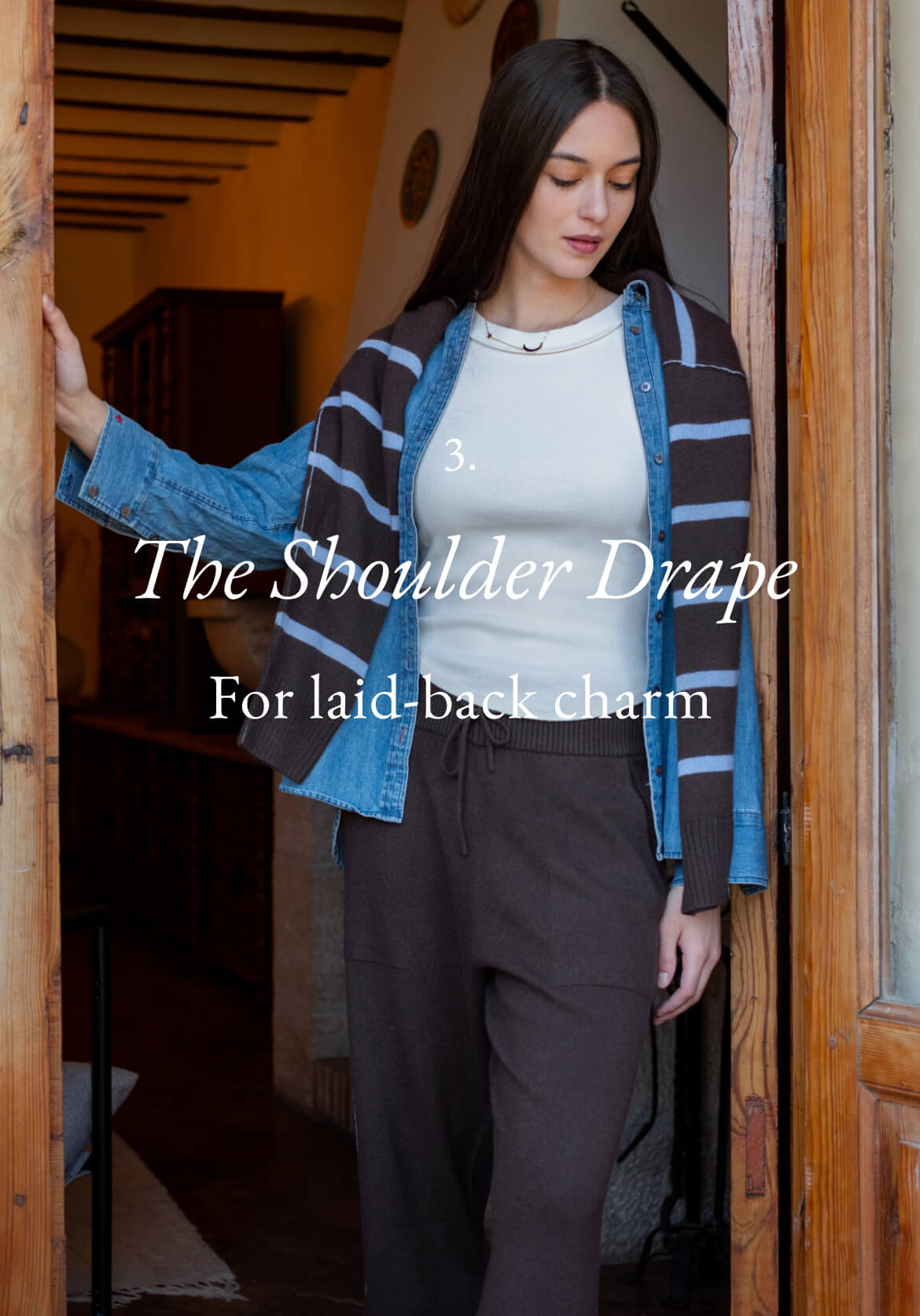 A woman wearing a striped cardigan over a white top and trousers, standing in a doorway. Text: 'The Shoulder Drape For laid-back charm'