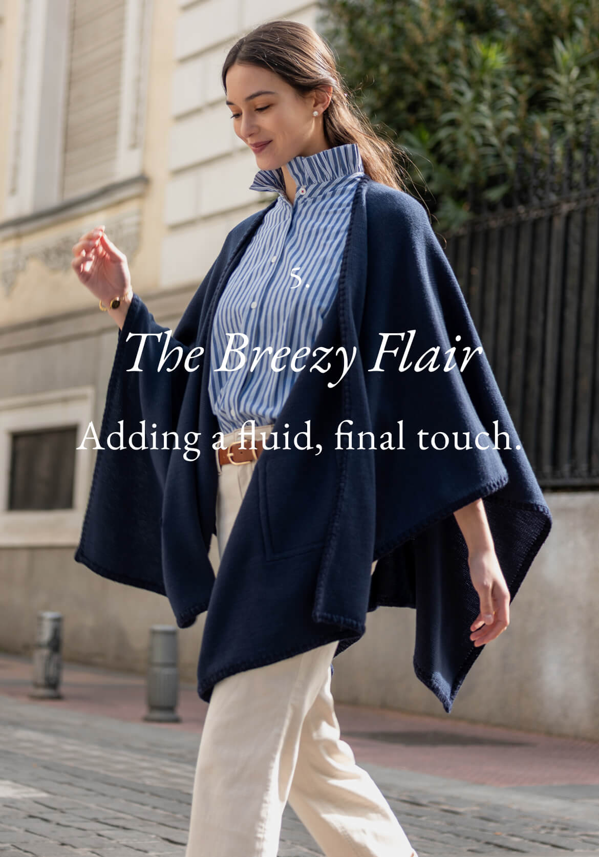 A woman wearing a navy blue knit wrap over a striped shirt and cream trousers, standing outdoors. Text: 'The Breezy Flair. Adding a fluid, final touch'