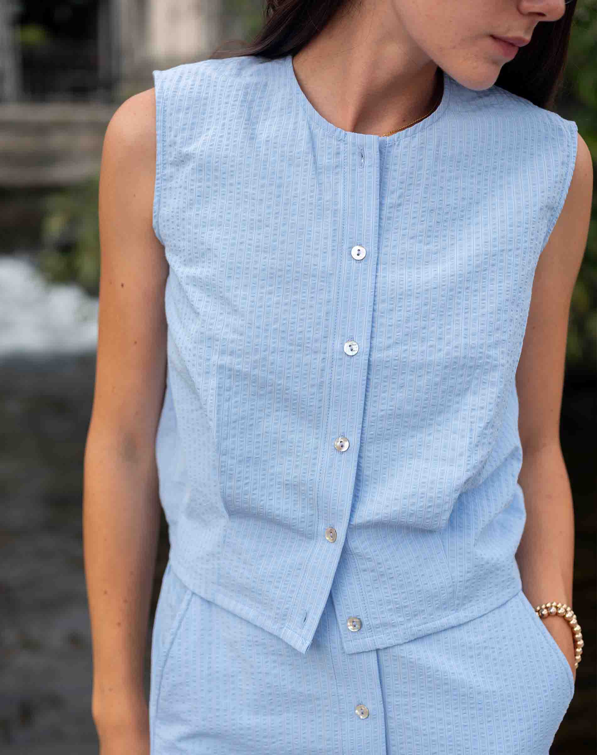 Person wearing a light blue sleeveless top with buttons, standing outdoors.