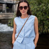 Woman wearing a light blue sleeveless top and skirt with sunglasses, standing outdoors near a water body.