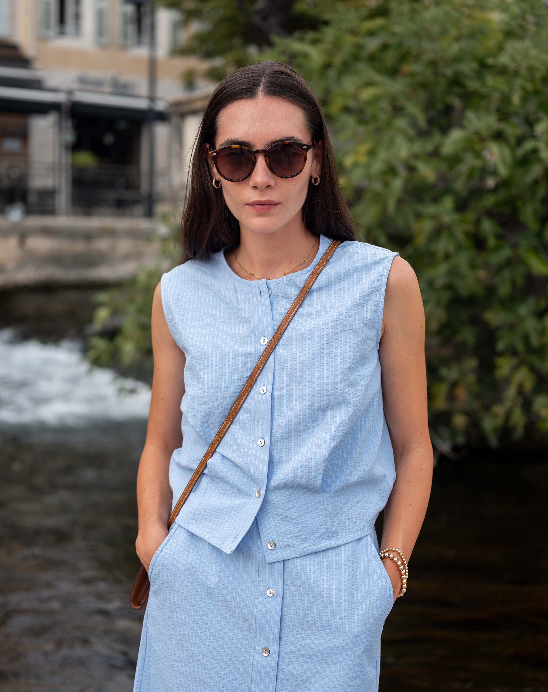 Woman wearing a light blue sleeveless top and skirt with sunglasses, standing outdoors near a water body.