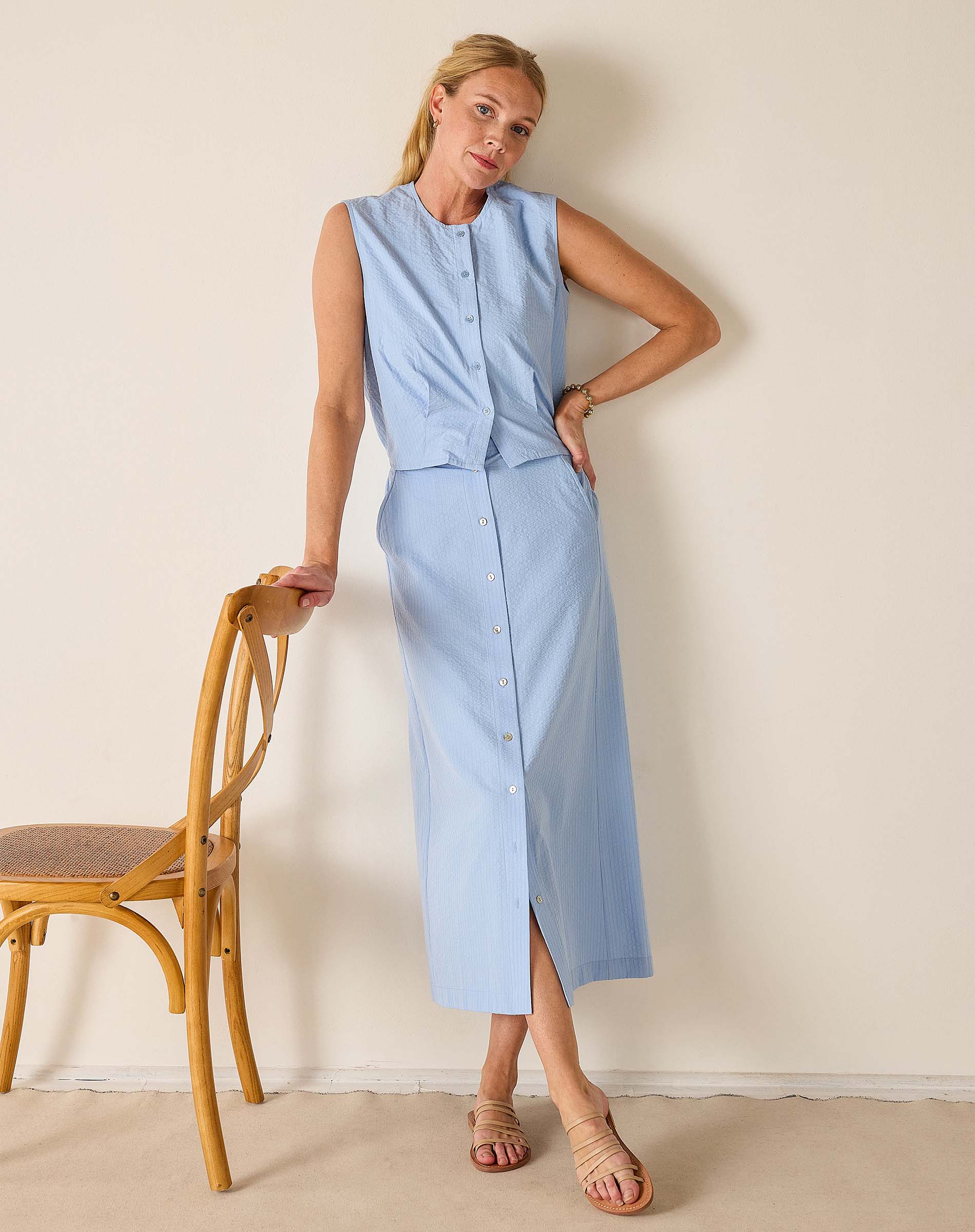 Woman wearing a light blue sleeveless dress standing next to a wooden chair against a plain wall.