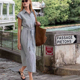 Woman in a gray dress standing by a river with a 'Passage Piétons' sign in the background.