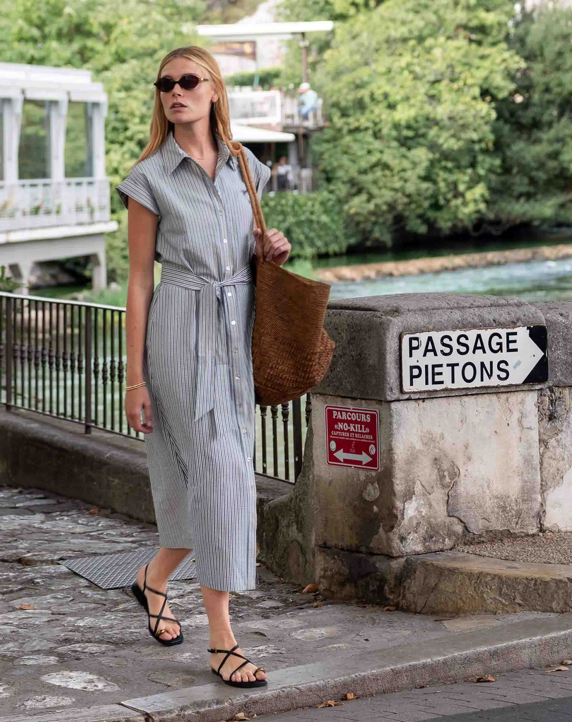 Woman in a gray dress standing by a river with a 'Passage Piétons' sign in the background.