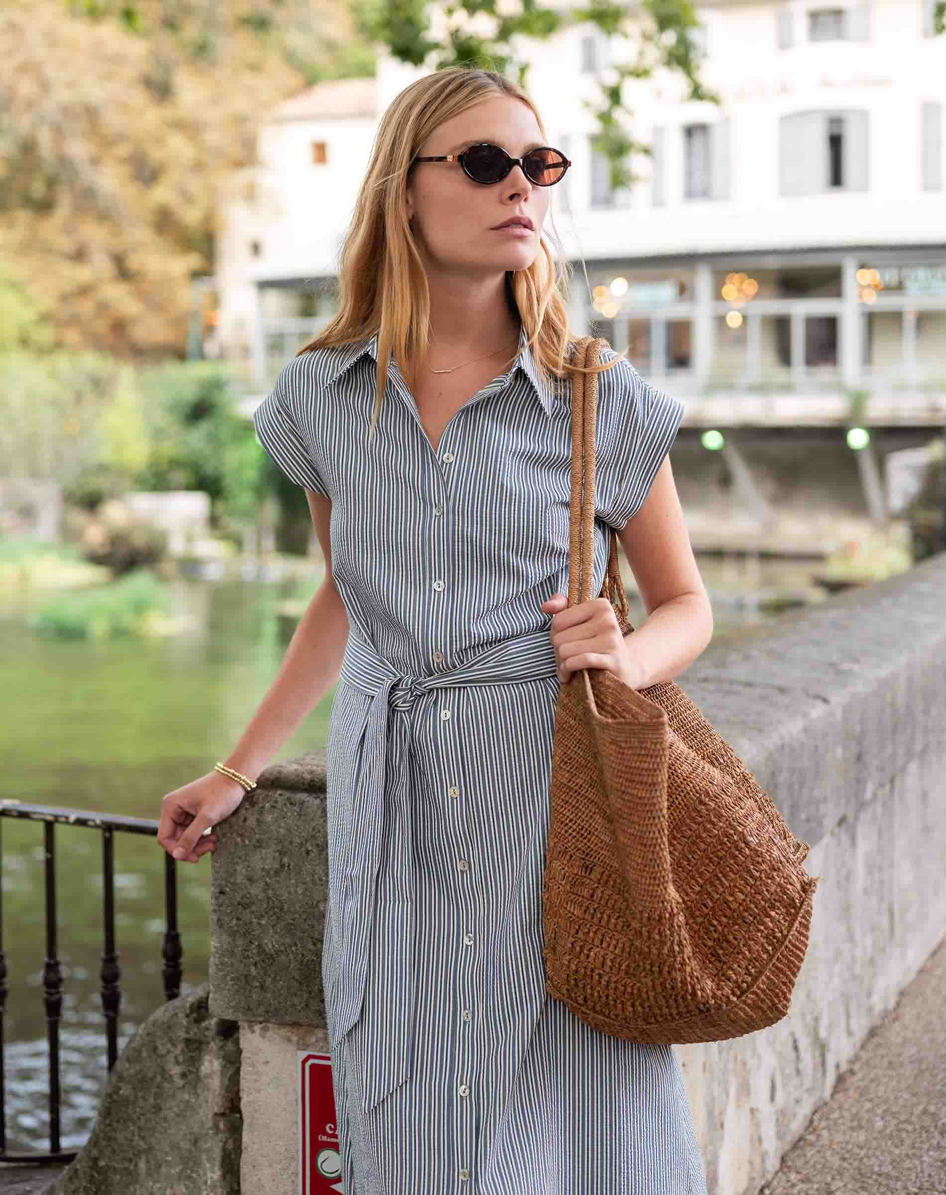 Woman in a striped dress holding a woven bag by a waterfront.