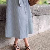 Person wearing a blue striped dress with black sandals standing against a stone wall.