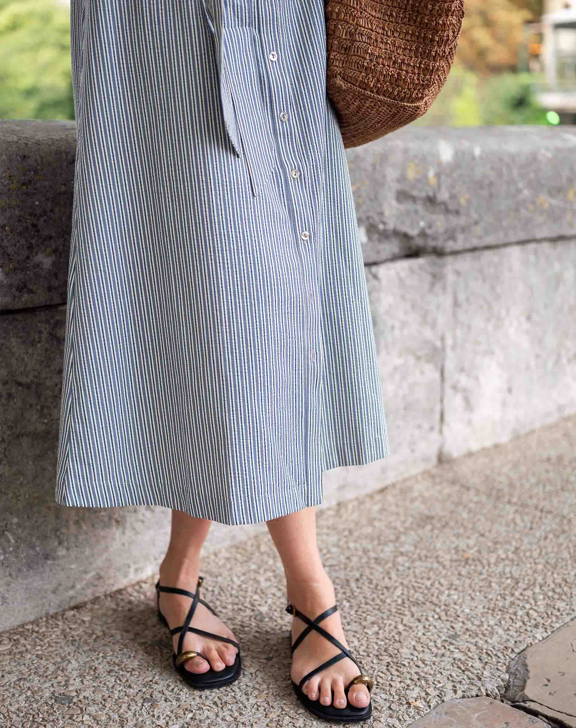 Person wearing a blue striped dress with black sandals standing against a stone wall.