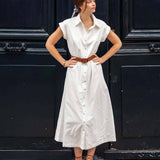 Woman wearing a white dress with a brown belt standing in front of a black door.