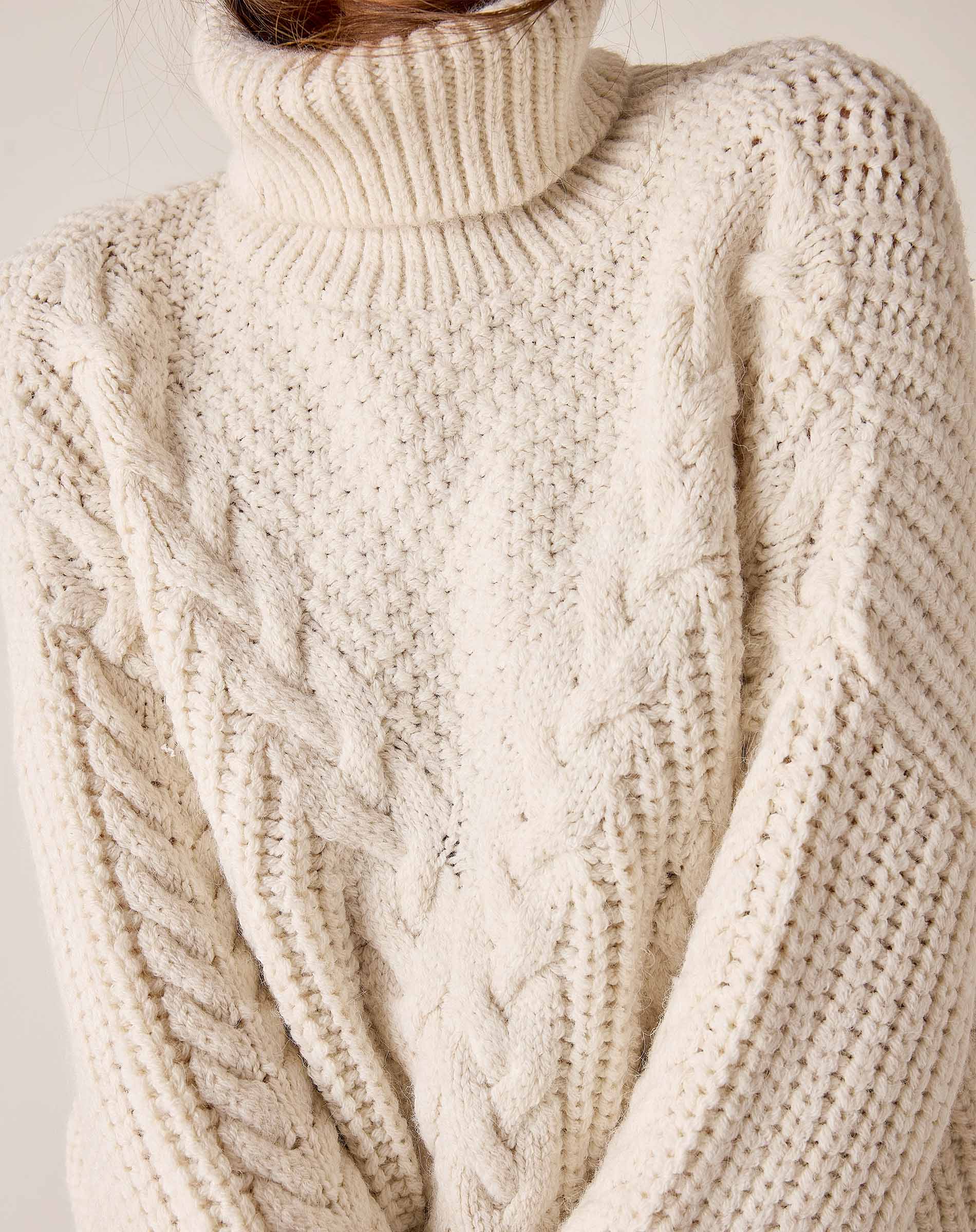 Cream cable knit turtleneck sweater with chunky ribbed neck and intricate stitch details.