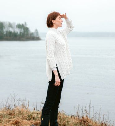 Red Hair female wearing a white sweater and black jeans looks at the horizon in front of the ocean. See our Maine collection.