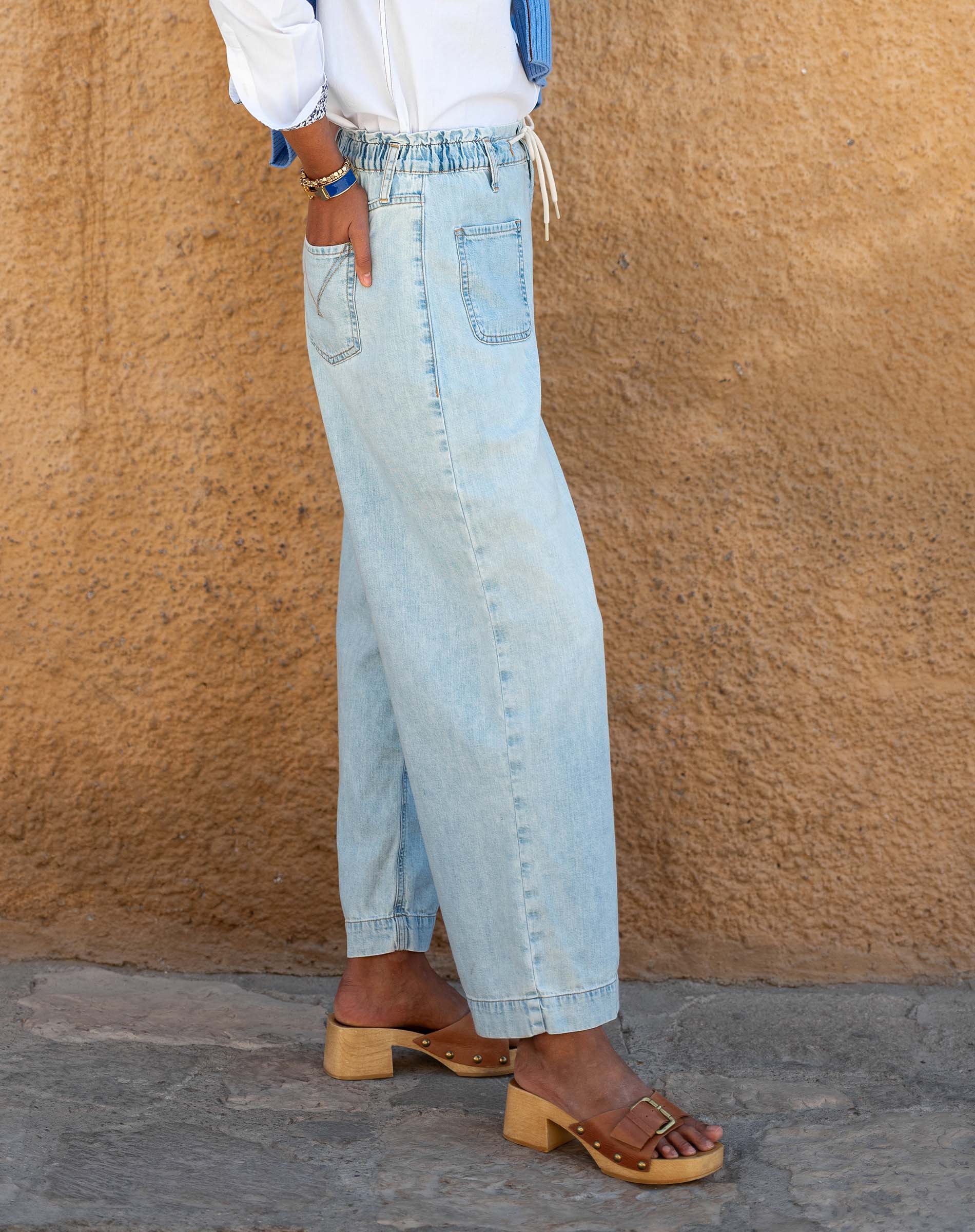Person wearing light blue jeans and wooden sandals against a beige wall.