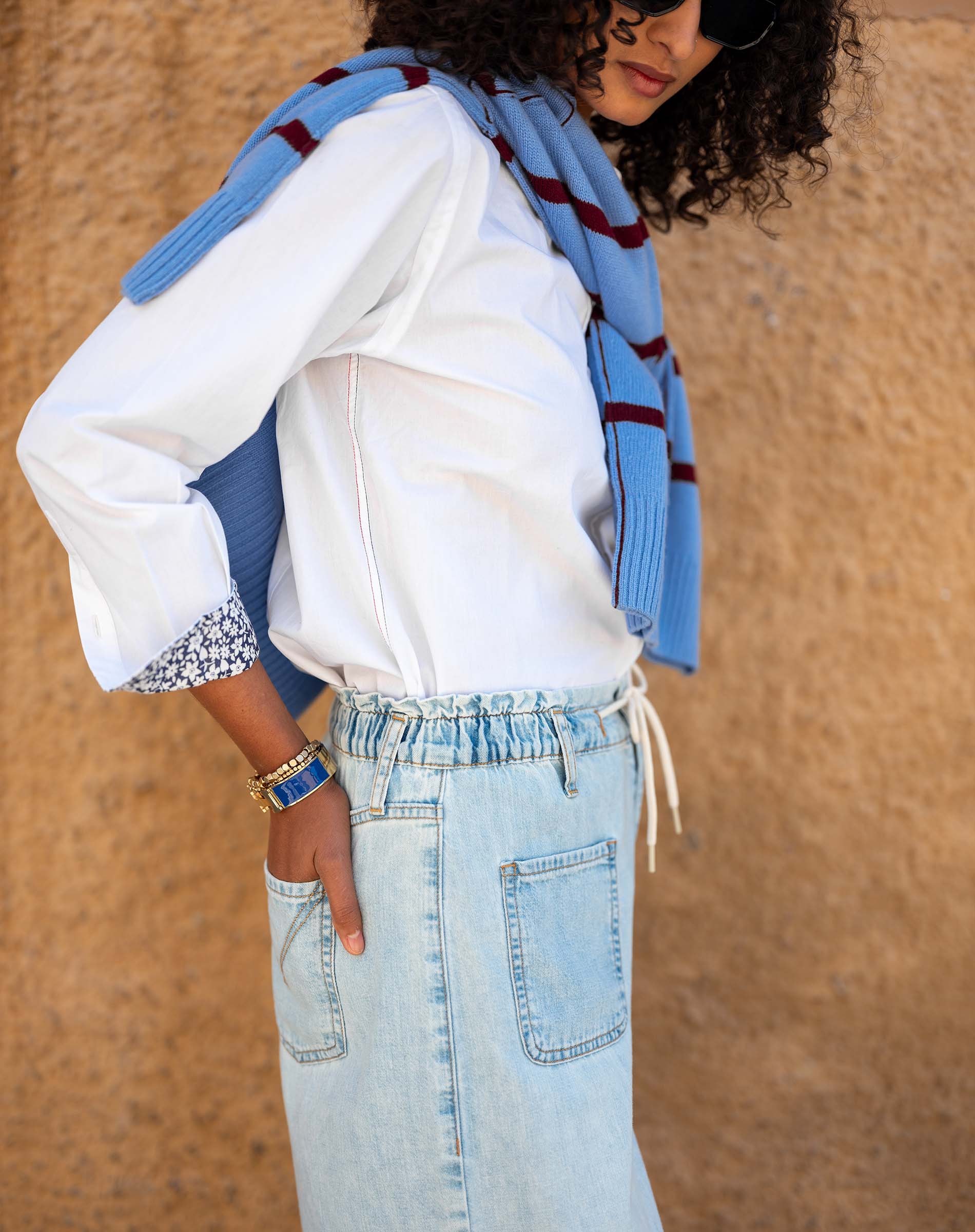 Person wearing a white shirt, blue jeans, and a blue scarf against a beige wall.