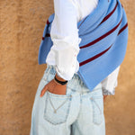 Person wearing a light blue cardigan with red stripes over a white shirt and light blue jeans, standing against a beige wall.