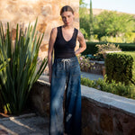Woman in black tank top and blue jeans standing outdoors near a stone wall and plants.