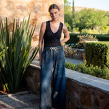 Woman in black tank top and blue jeans standing outdoors near a stone wall and plants.