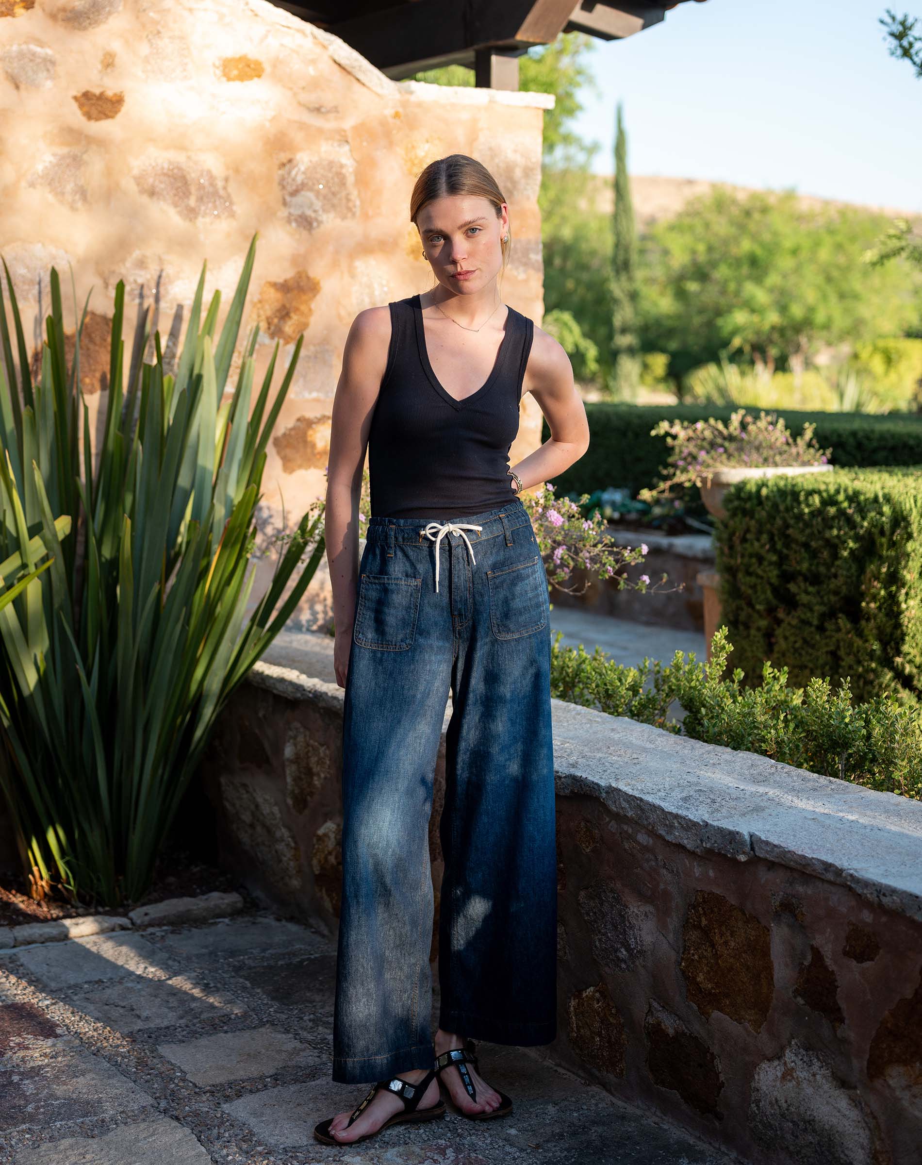 Woman in black tank top and blue jeans standing outdoors near a stone wall and plants.