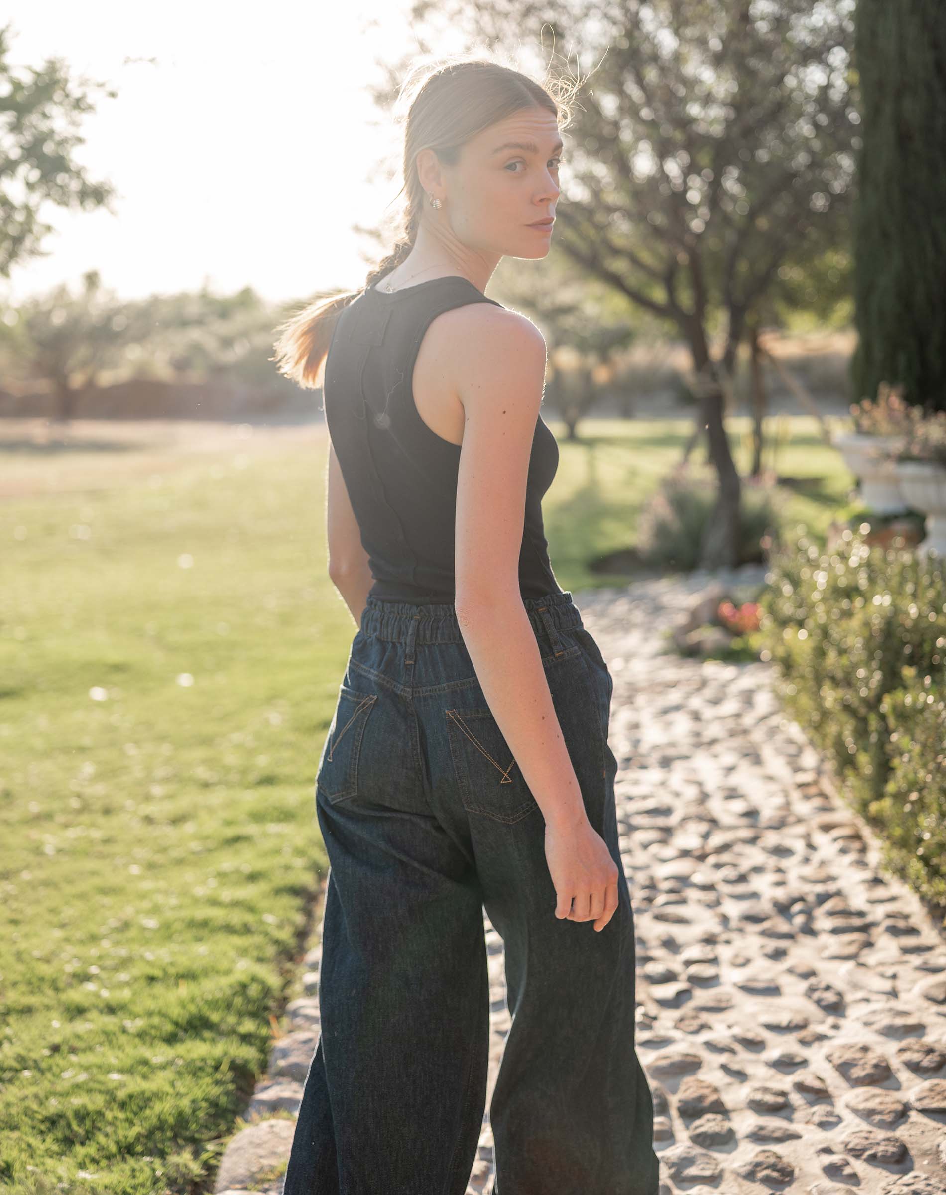 Woman in a black sleeveless top and dark blue jeans walking on a stone path in a garden.