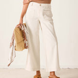 Person wearing white pants and brown sandals holding a woven bag against a plain background