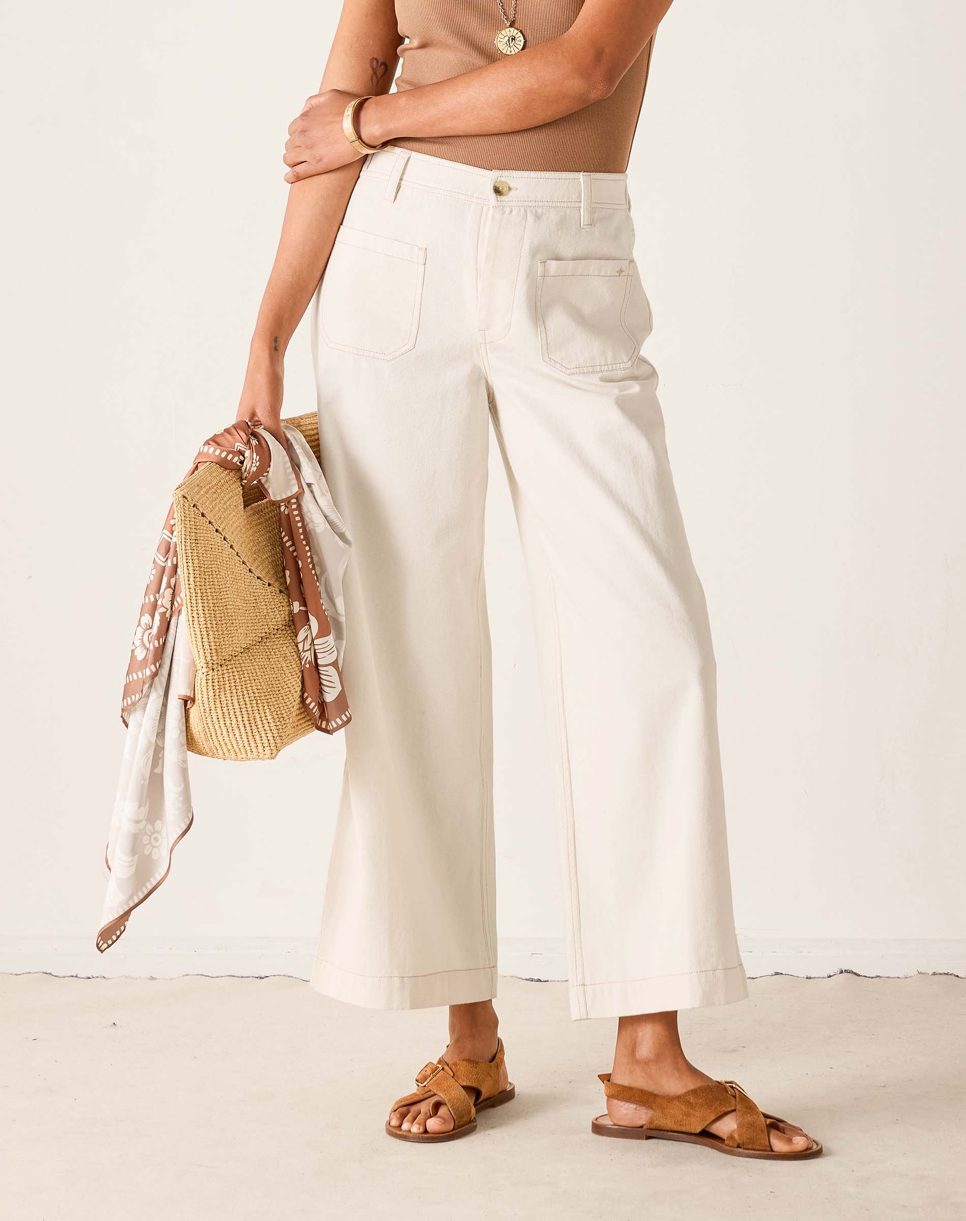Person wearing white pants and brown sandals holding a woven bag against a plain background