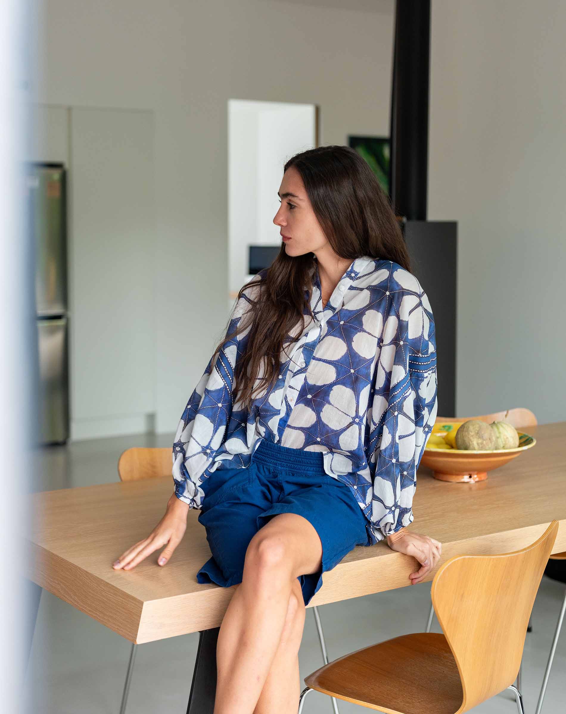 Woman sitting at a kitchen table wearing a blue and white floral shirt and shorts.