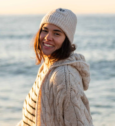 female wearing neutral beanie with black stripe sweater and neutral duster on the beach