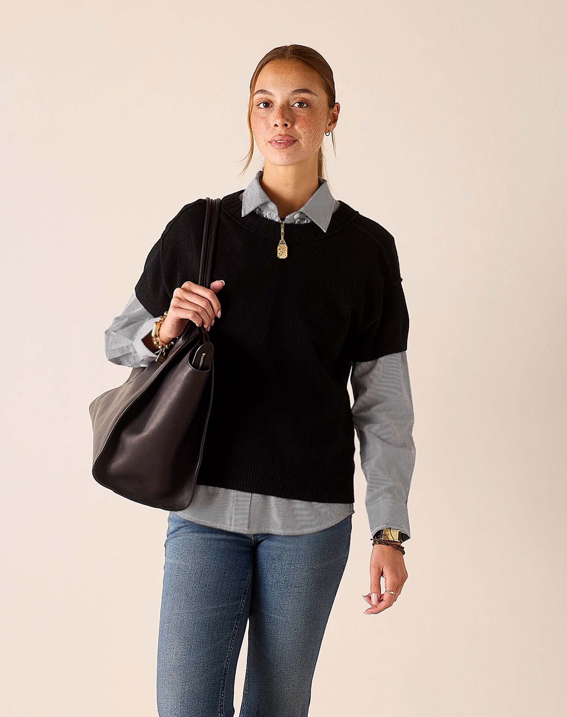 Woman in a black cashmere crewneck sweater over a grey button-down shirt and blue jeans.