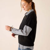 A woman in a black cashmere crewneck sweater over a striped button-up shirt and denim jeans.