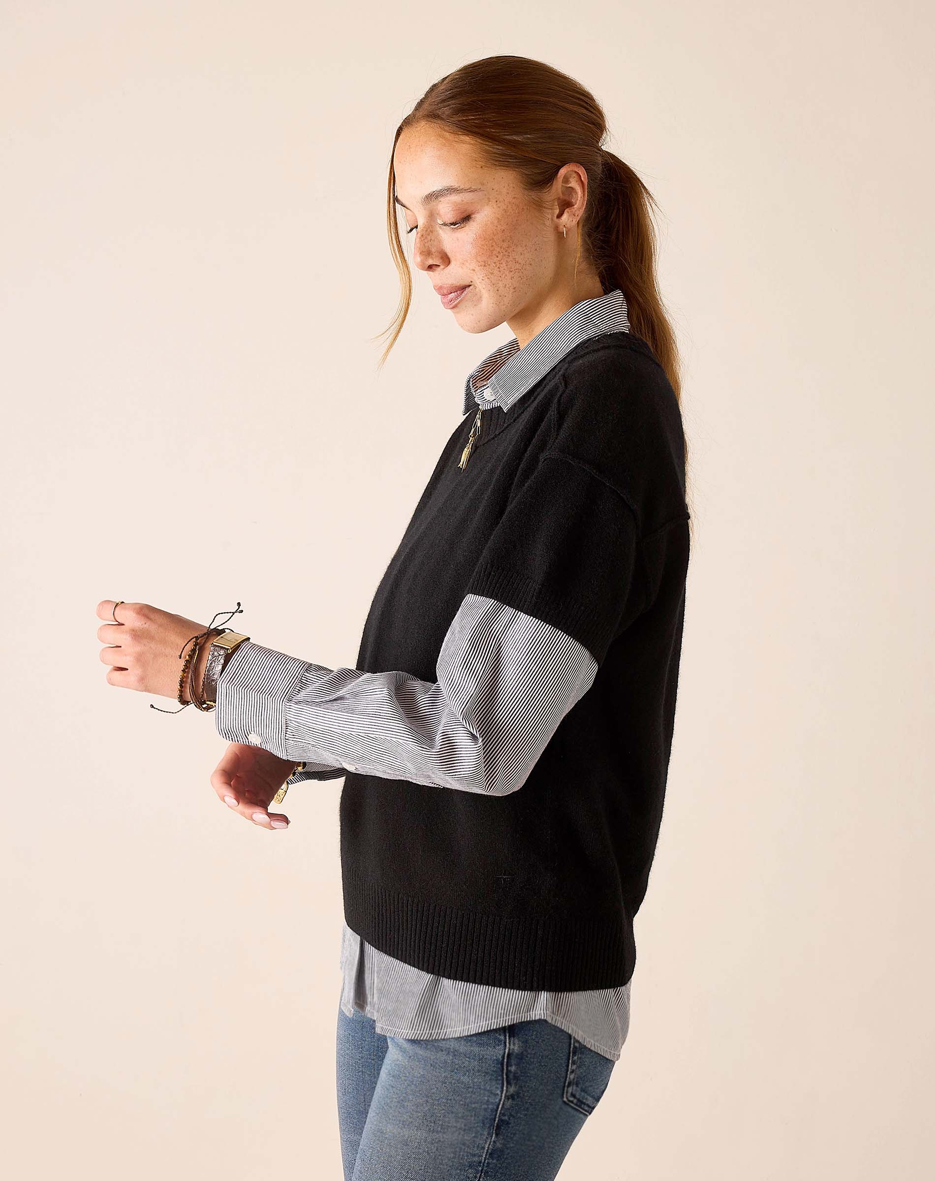A woman in a black cashmere crewneck sweater over a striped button-up shirt and denim jeans.