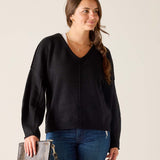 Woman wearing a black sweater and blue jeans, holding a clutch against a plain background