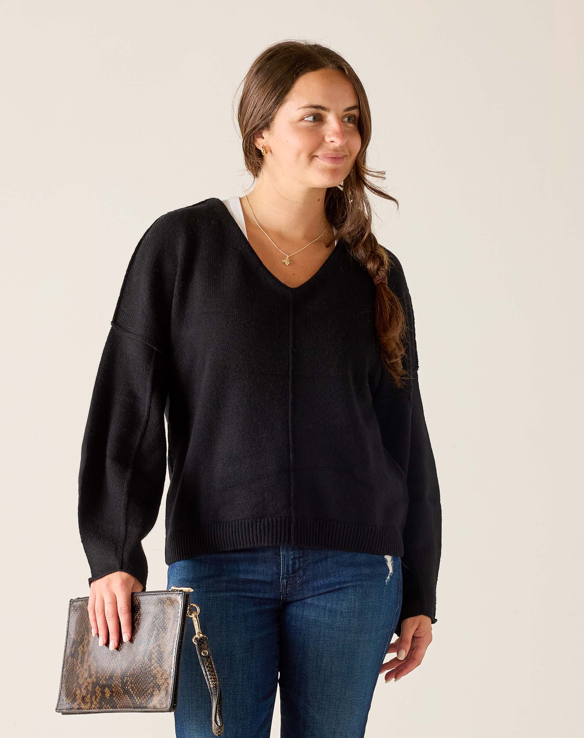 Woman wearing a black sweater and blue jeans, holding a clutch against a plain background