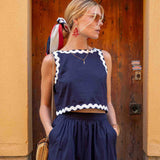 Woman wearing a navy blue sleeveless top with white trim and dark blue pants, standing in front of a wooden door.