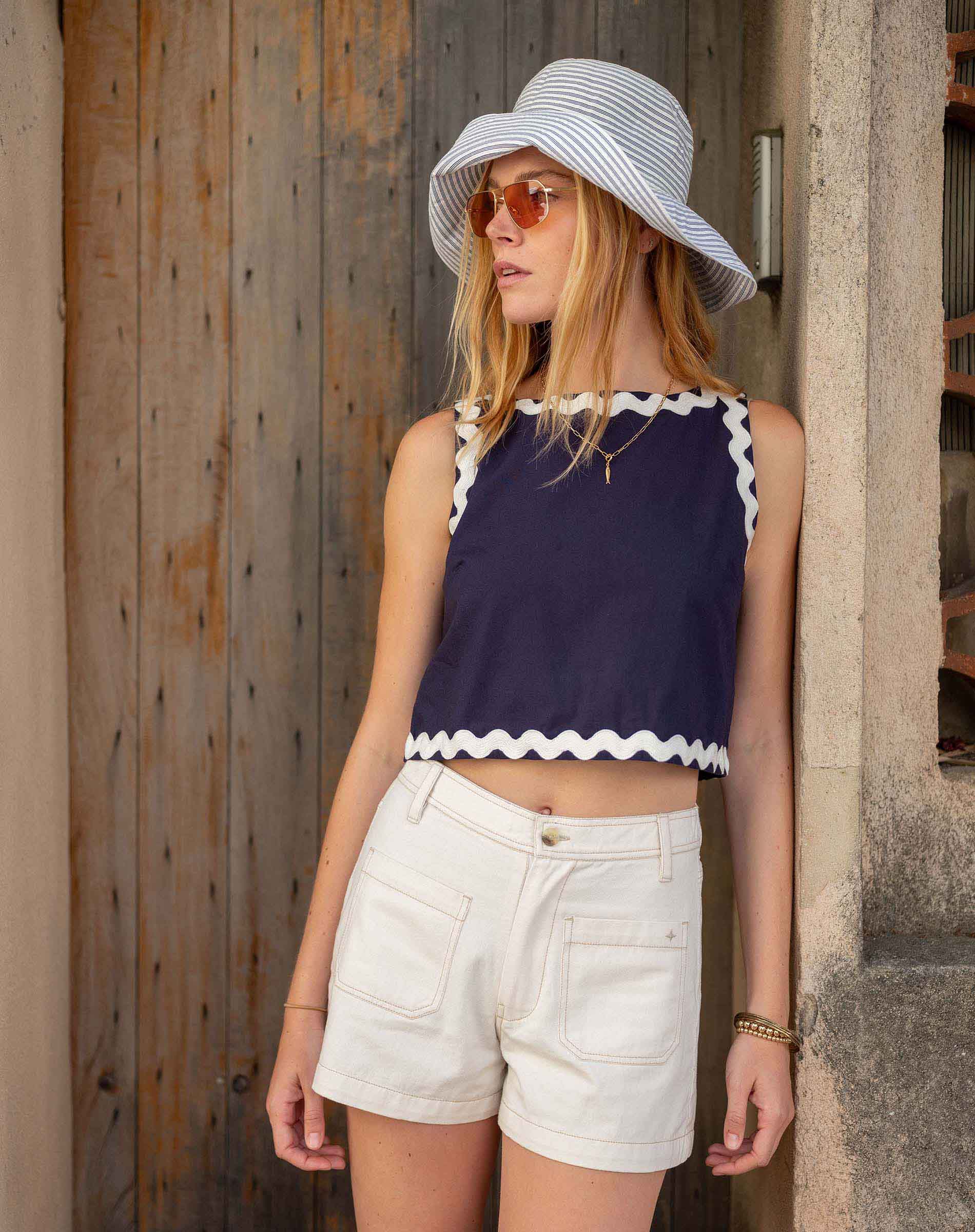 Woman wearing a navy top with white trim and white shorts, standing against a wooden wall.