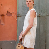 Woman in a white dress holding a woven bag against a textured wall.