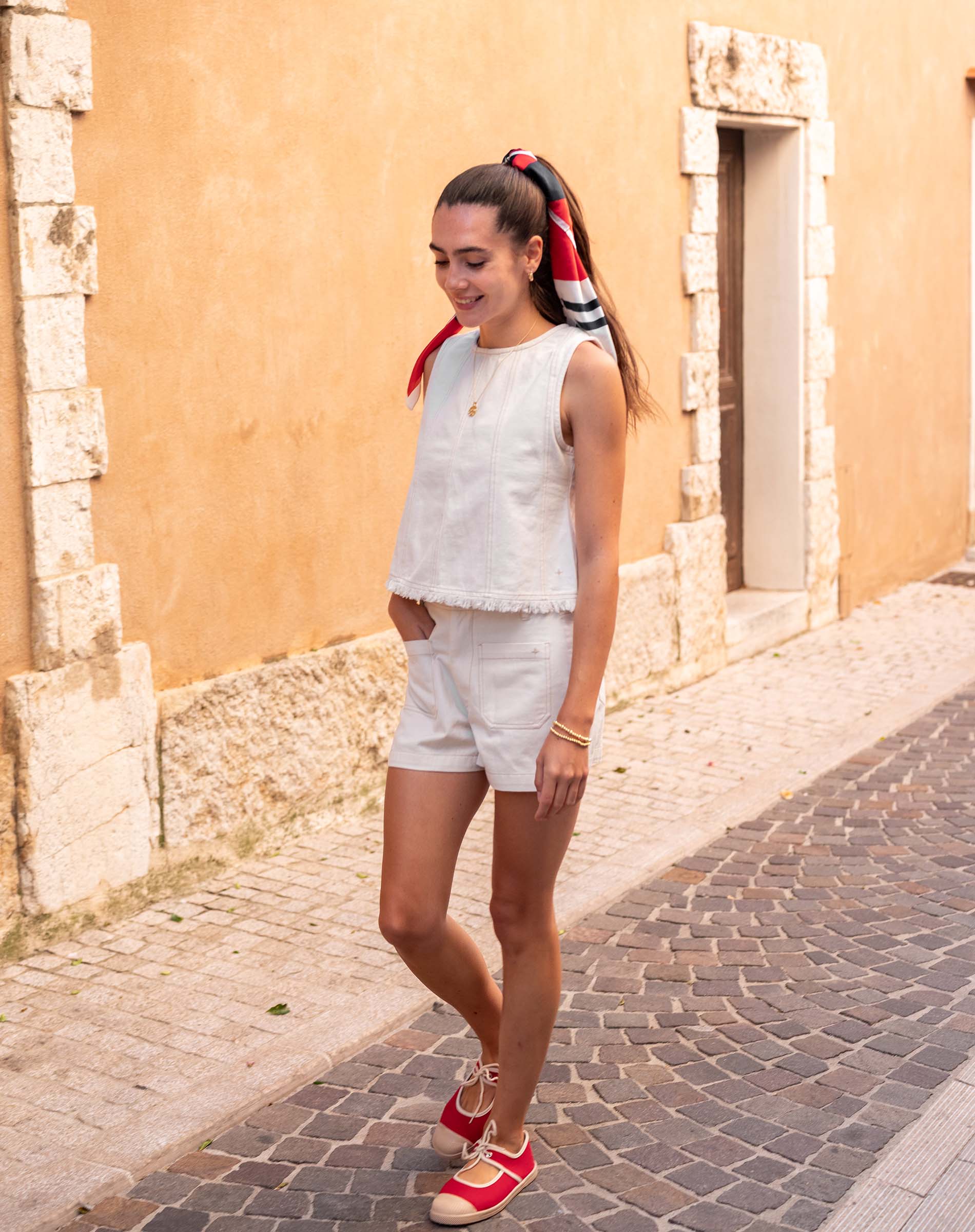 Woman walking on a cobblestone street wearing a white sleeveless top and shorts with red shoes.