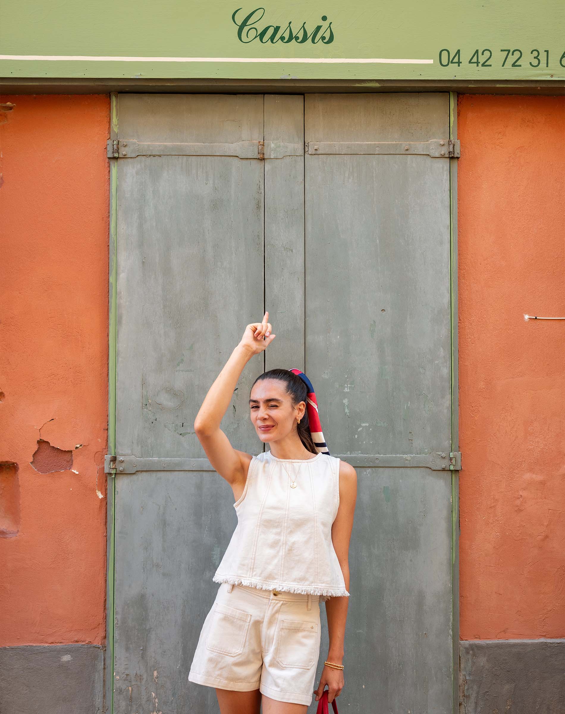 Woman in a white outfit posing in front of a green and orange building with 'Cassis' written on it.
