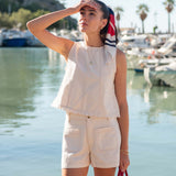 Woman in a white sleeveless top and shorts standing by a waterfront with boats in the background.