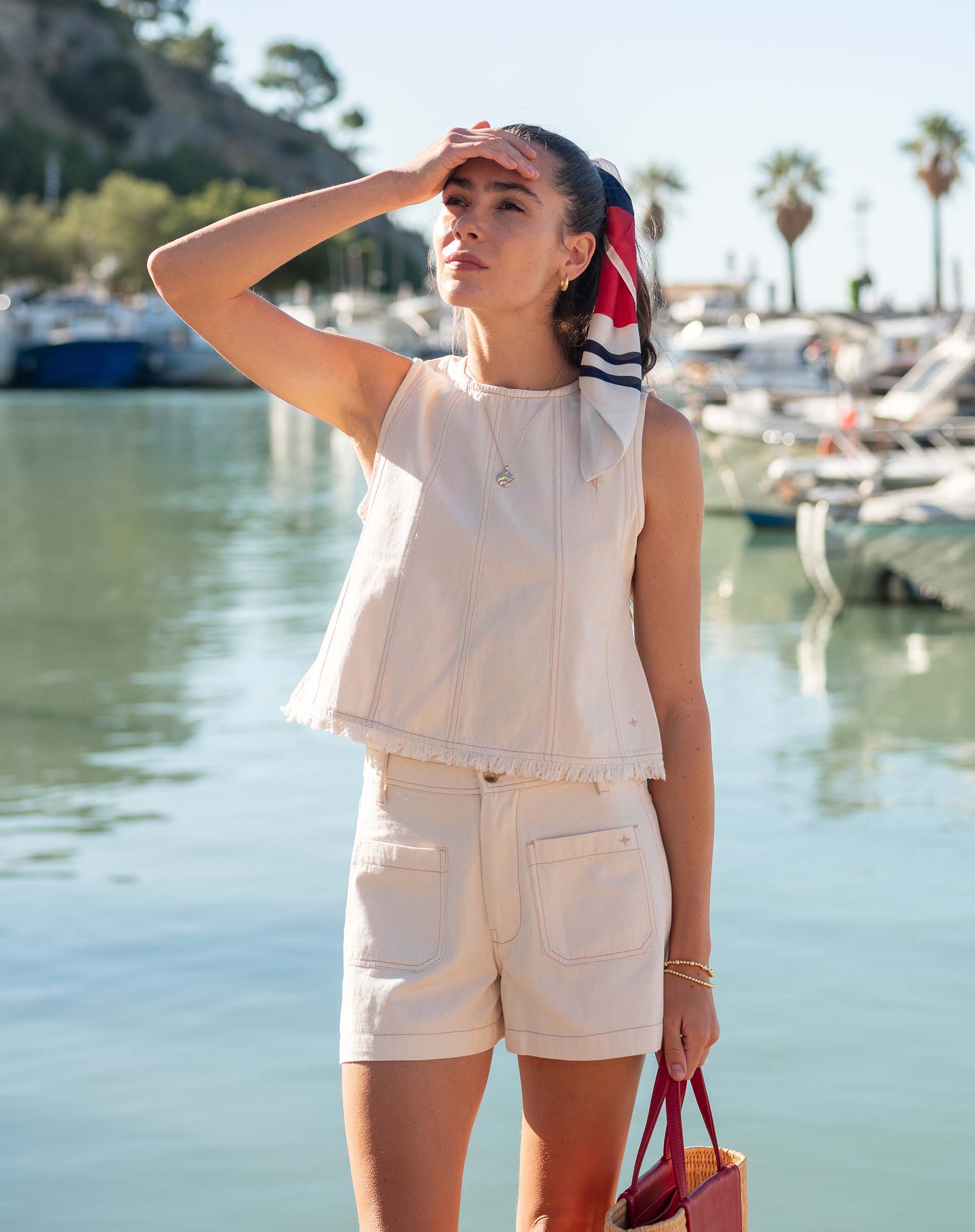 Woman in a white sleeveless top and shorts standing by a waterfront with boats in the background.
