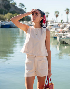 Woman in a white sleeveless top and shorts standing by a waterfront with boats in the background.