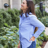 Woman wearing a blue striped shirt and jeans standing outdoors with greenery in the background