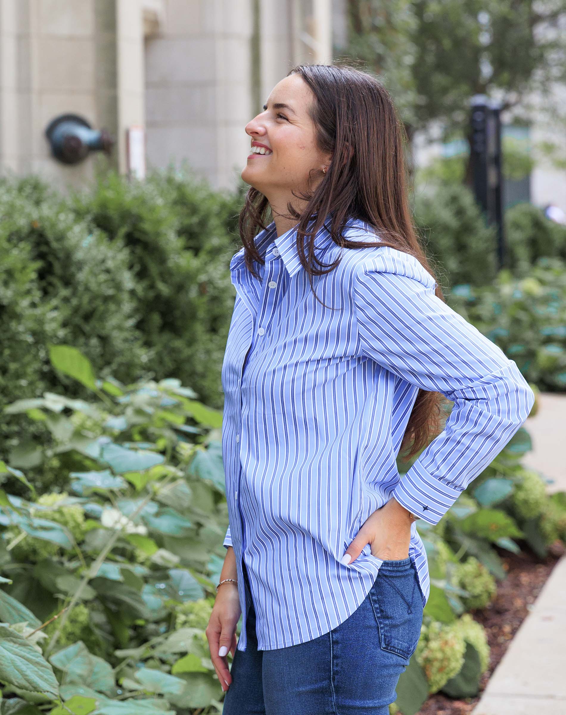 Woman wearing a blue striped shirt and jeans standing outdoors with greenery in the background