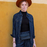 Person wearing a denim jacket, black turtleneck, and brown hat against a yellow wall