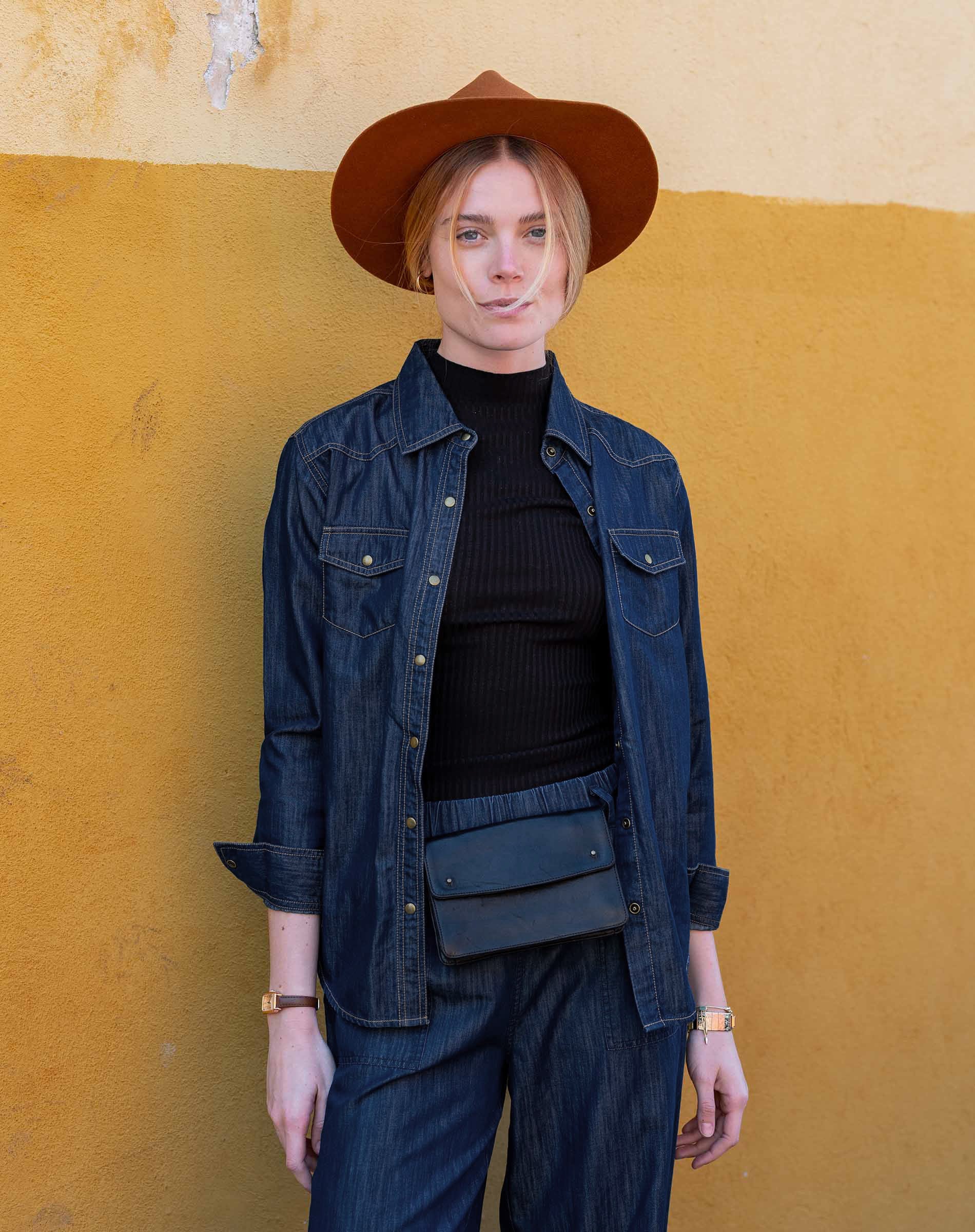 Person wearing a denim jacket, black turtleneck, and brown hat against a yellow wall