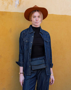 Person wearing a denim jacket, black turtleneck, and brown hat against a yellow wall