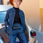 Person wearing a blue denim outfit and brown hat leaning against a vintage car.