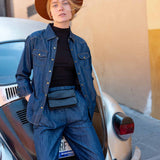 Person wearing a blue denim outfit and brown hat leaning against a vintage car.