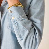 Light blue denim shirt with rolled-up sleeve and gold bracelet on a neutral background