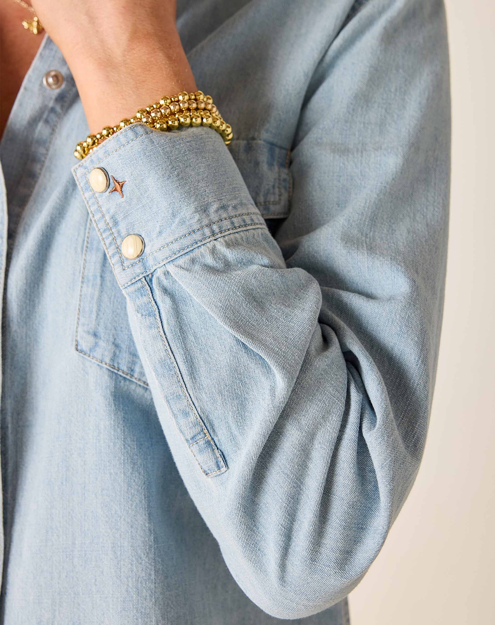 Light blue denim shirt with rolled-up sleeve and gold bracelet on a neutral background