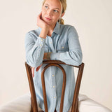 Woman sitting on a wooden chair wearing a light blue denim shirt against a plain background
